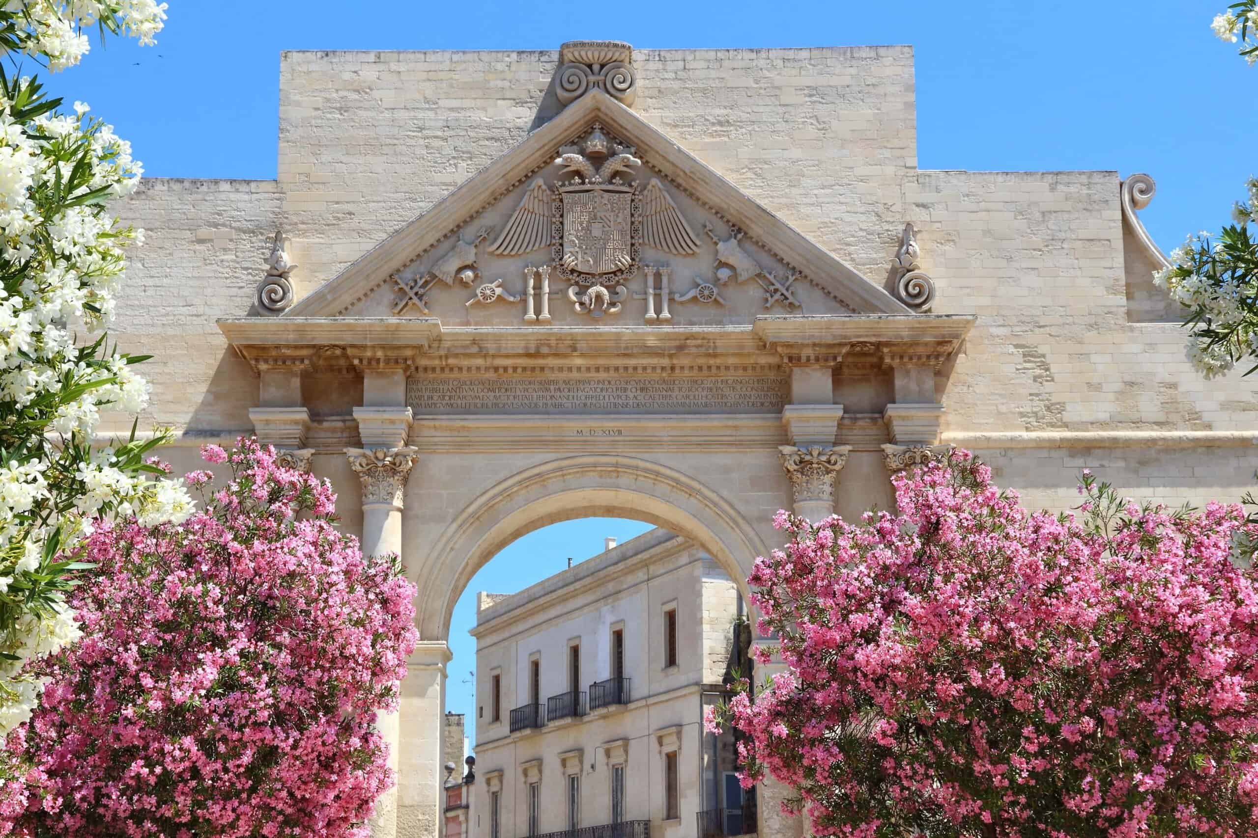 Gates of the city of Lecce framed by two trees with pink flowers in bloom.